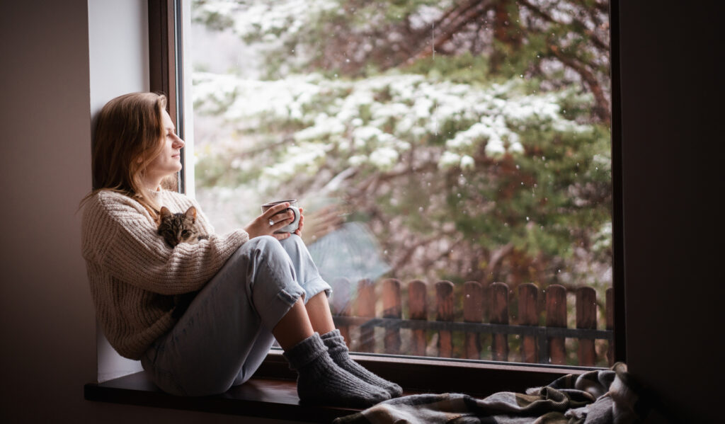 A young woman sitting comfortably on a ledge next to a large bay window in her home. Snow can be seen on the tree limbs in the background. She is wearing a sweater, jeans and heavy socks. She has a cup of coffee or tea in her hands and her cat is resting in her lap.